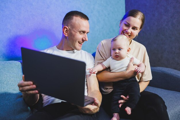 Eine junge Mutter und ein junger Vater sitzen mit ihrem neugeborenen Sohn auf der Couch und sehen sich einen Film auf einem Laptop an Familienbeziehungen mit einem kleinen Baby Ein Familienpaar mit einem Kind zu Hause auf dem Sofa