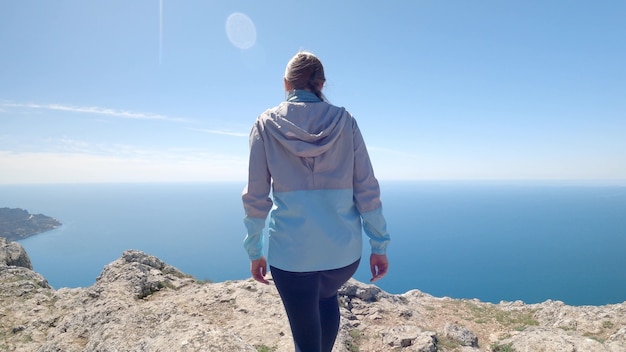 Eine junge Reisende kletterte auf die höchste Klippe mit Blick auf den endlosen Ozean. Sie breitet die Arme seitlich aus und genießt die schöne Aussicht. Ruhige und atemberaubende Landschaft. 4K UHD