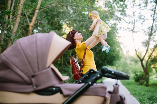 Eine junge rothaarige Frau in einem gelben Hemd geht mit einem kleinen Baby in einem Kinderwagen an einem hellen Sommertag im Park spazieren