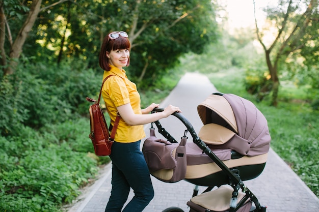 Eine junge rothaarige Frau in einem gelben Hemd geht mit einem kleinen Baby in einem Kinderwagen an einem hellen Sommertag im Park spazieren