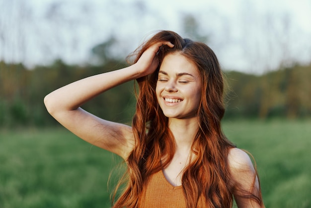 Eine junge rothaarige Frau lächelt im Sonnenuntergang in einem grünen Park und hält sich eine Hand ans Haar