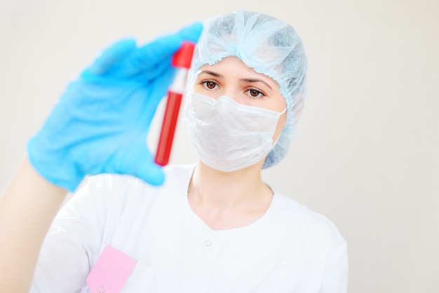 Eine junge Ärztin oder Krankenschwester in einer medizinischen Maske und Gummihandschuhen, die ein Reagenzglas mit einer Blutprobe hält