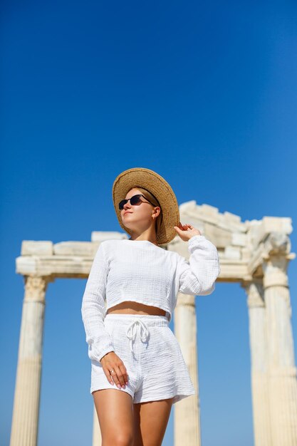 Eine junge, schlanke, gebräunte Frau in weißen Shorts und einem T-Shirt mit Sonnenbrille und Hut Gehen Sie an einem heißen sonnigen Tag spazieren