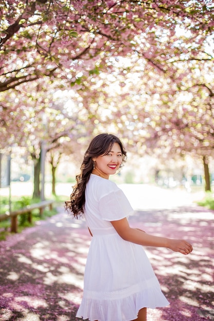 Eine junge schöne asiatische Frau in einem weißen Kleid geht in einem Blumenpark. Sakura. Blühende Bäume. Frühling.