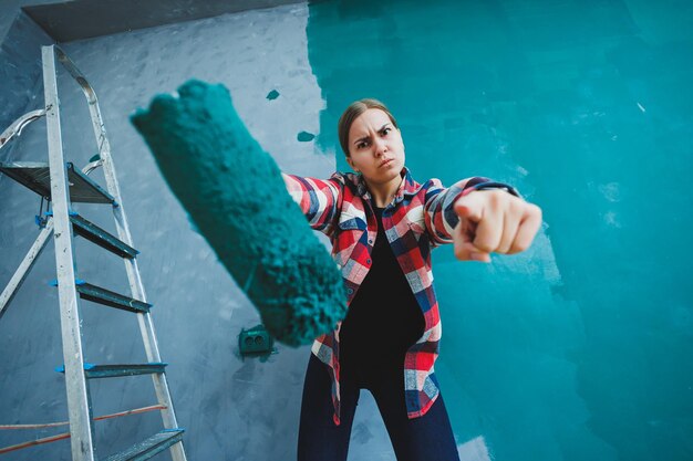 Eine junge schöne Designerin macht Reparaturen malt eine graue Wand mit grüner Farbe mit einer Walze Kariertes Hemd lange Haare und Jeans