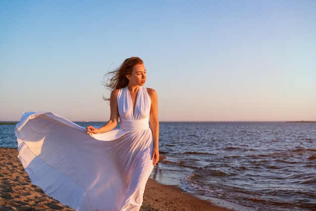 Eine junge schöne erwachsene brünette Frau in einem weißen flatternden Kleid steht am sandigen Ufer des ...