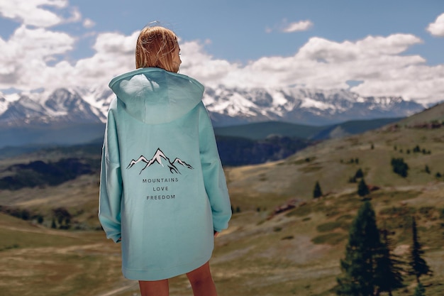 Eine junge schöne Frau in einem blauen, übergroßen Hoodie steht mit dem Rücken und blickt auf die malerische, fabelhafte Landschaft der Rocky Mountains.