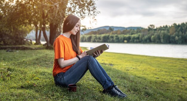 Eine junge schöne Frau in hellen Kleidern sitzt auf dem grünen Gras und genießt das Lesen eines Buches in der Natur. Konzept eines angenehmen Zeitvertreibs, Ruhe, Entspannung.