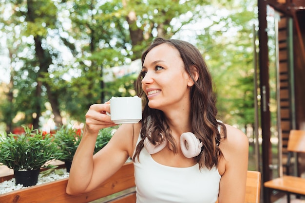 Eine junge schöne Frau mit Kopfhörern sitzt an einem Tisch in einem Sommercafé und trinkt Kaffee oder Tee Generation z