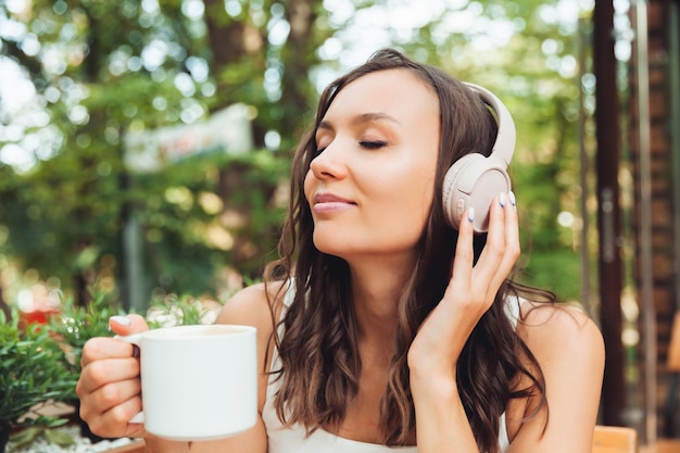 Eine junge schöne Frau mit Kopfhörern sitzt an einem Tisch in einem Sommercafé und trinkt Kaffee oder Tee Generation z