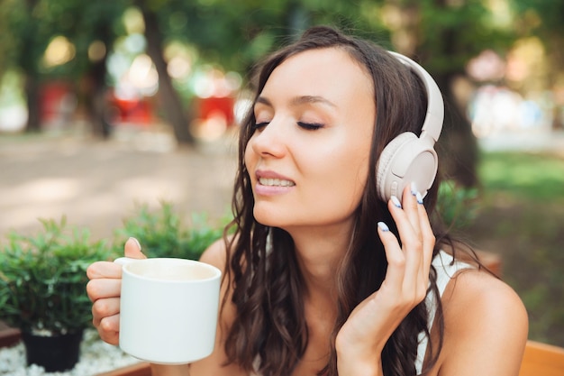 Eine junge schöne Frau mit Kopfhörern sitzt an einem Tisch in einem Sommercafé und trinkt Kaffee oder Tee Generation z