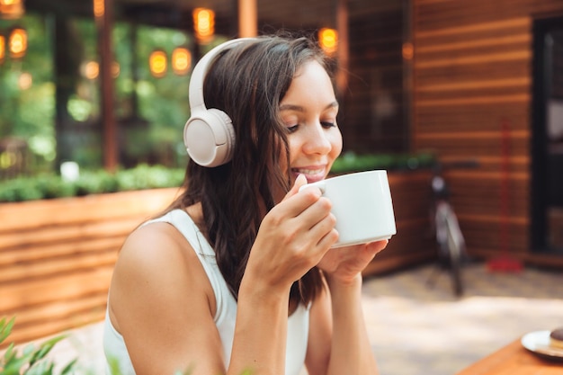 Eine junge schöne Frau mit Kopfhörern sitzt an einem Tisch in einem Sommercafé und trinkt Kaffee oder Tee Generation z