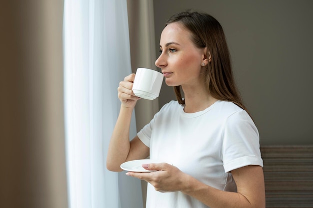 Eine junge schöne Frau schaut durch das Fenster und trinkt morgens Kaffee