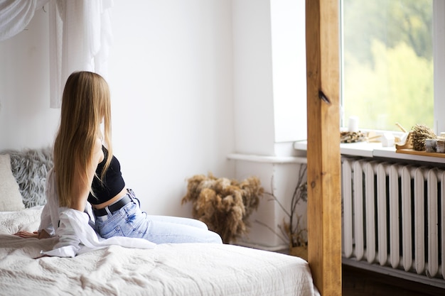 Eine junge schöne Frau sitzt auf dem Bett und schaut aus dem Fenster