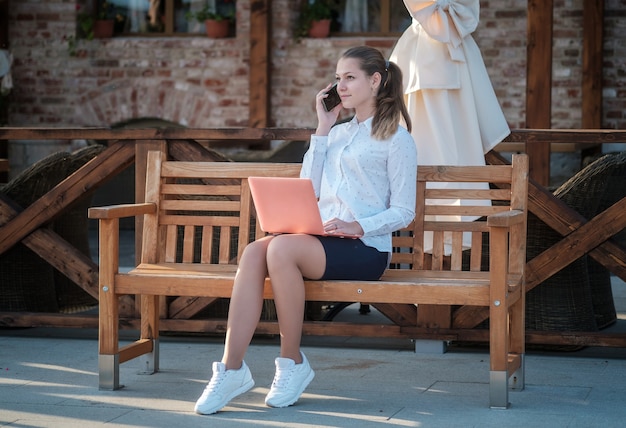 Eine junge schöne Frau sitzt auf einer Bank und hält ein Smartphone und einen Laptop. Student mit einem Laptop und Smartphone auf der Stadtstraße