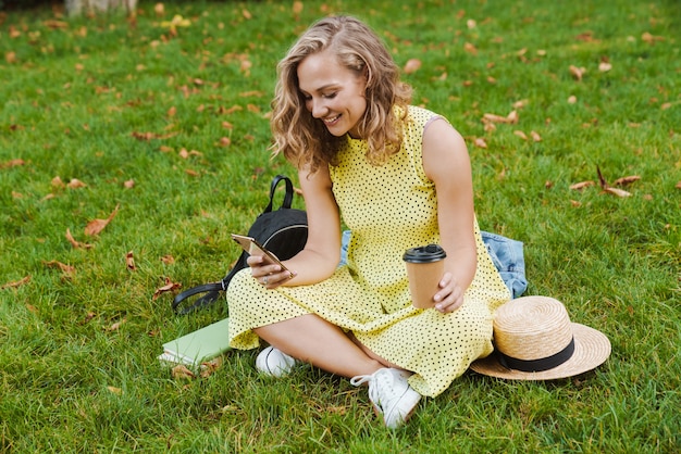 eine junge schöne fröhliche frau sitzt auf gras draußen im naturpark und hält kaffee mit handy.