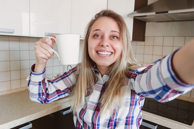 Eine junge schöne kaukasische lächelnde blonde Frau im karierten Hemd mit einer weißen Tasse Kaffee oder Tee nimmt ein Selfie