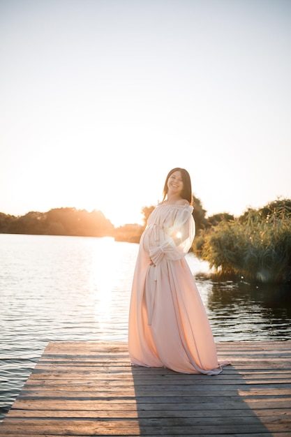 Eine junge schwangere Frau in einem Chiffonkleid steht auf einem Pier am Fluss vor dem Hintergrund eines orangefarbenen Sonnenuntergangs