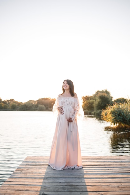 Eine junge schwangere Frau in einem Chiffonkleid steht auf einem Pier am Fluss vor dem Hintergrund eines orangefarbenen Sonnenuntergangs