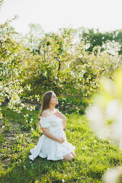 Eine junge schwangere Frau in einem weißen Kleid geht durch einen blühenden Apfelgarten. Glückliche schwangere Frau, glückliche zukünftige Mutter