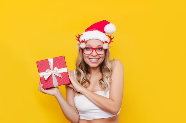 Eine junge sexy Frau in Weihnachtsbrille mit Hirschgeweih zeigt eine Geschenkbox auf gelbem Hintergrund