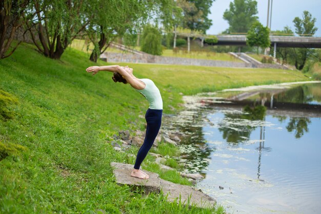 Eine junge Sportlerin übt Yoga auf einem grünen Rasen am Fluss, Yoga assans Haltung. Meditation und Einheit mit der Natur