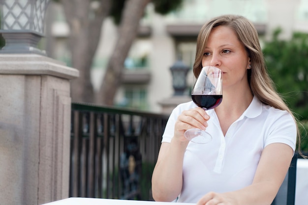 Eine junge stilvolle schöne blonde Frau in einem weißen Hemd trinkt am Abend Rotwein auf der offenen Sommerveranda des Vodino-Restaurants. Ein schöner Look und ein leichtes verspieltes Lächeln