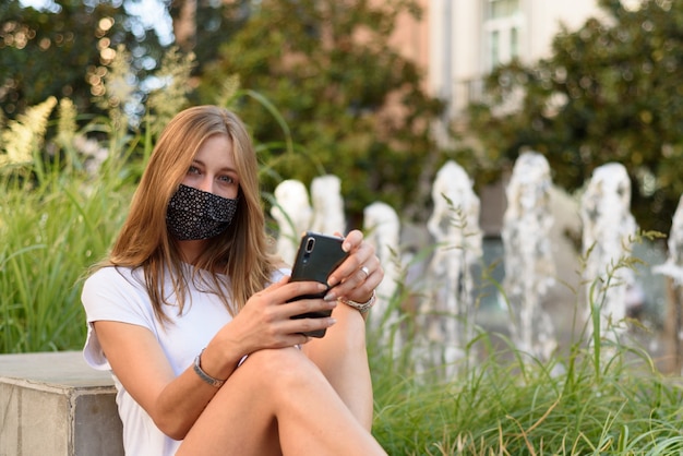 Eine junge weiße Frau mit einer Maske, die durch ihr Telefon auf einer Straße schaut