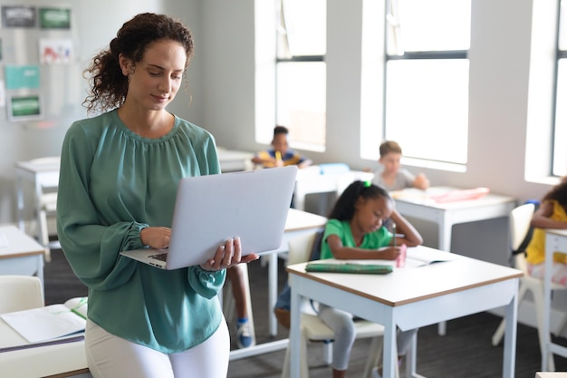 Eine junge weiße Lehrerin benutzt einen Laptop mit multiracialen Grundschülern im Hintergrund