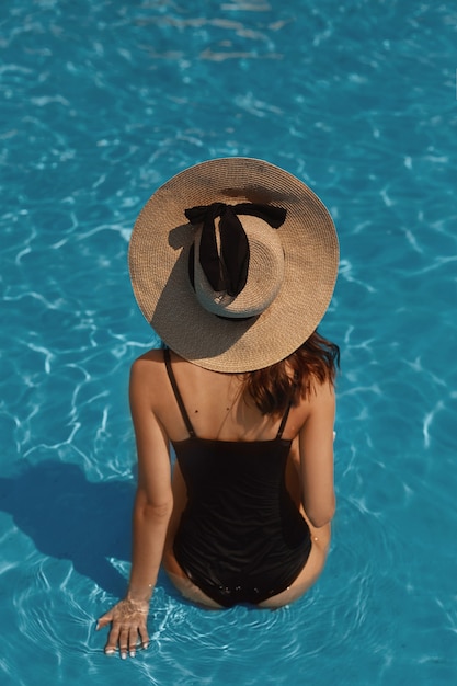 Eine junge wunderschöne Frau mit sexy großen Brüsten und schlanker Taille in einem schwarzen Badeanzug, der im Schwimmbad draußen an einem Sommertag aufwirft.