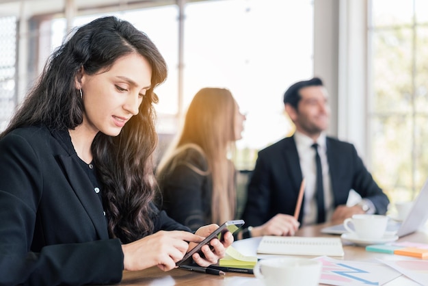 Eine kaukasische Managerin in einem schwarzen Anzug, die auf einem Smartphone sitzt und arbeitet, eine Geschäftsfrau, die auf einem Touchscreen des Mobiltelefons tippt, während sie sich im Büro mit unscharfen Kollegen im Hintergrund trifft.