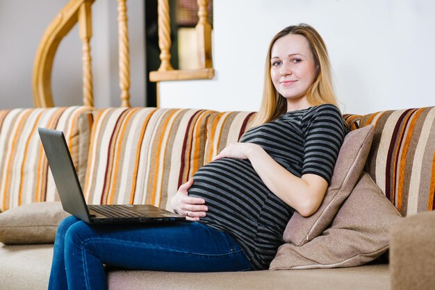 Eine kaukasische schwangere Frau, die mit einem Laptop auf dem Sofa sitzt