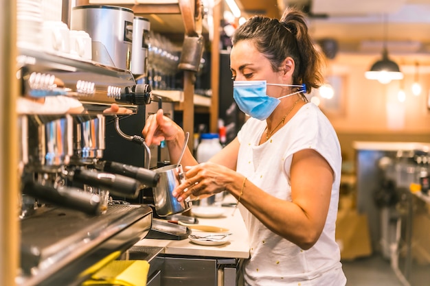 Eine Kellnerin mit einer Gesichtsmaske in einer Bar. An einem Frühlingsmorgen mit der Kaffeemaschine einen Kaffee mit Milch zubereiten