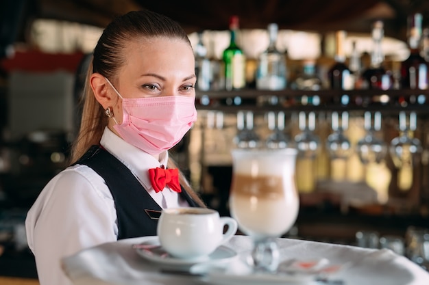 Eine Kellnerin mit europäischem Aussehen in einer medizinischen Maske serviert Latte-Kaffee.