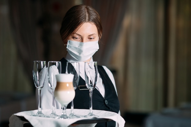 Eine Kellnerin mit europäischem Aussehen in einer medizinischen Maske serviert Latte-Kaffee.