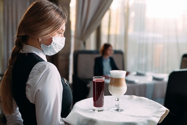 Eine Kellnerin mit europäischem Aussehen in einer medizinischen Maske serviert Latte-Kaffee.