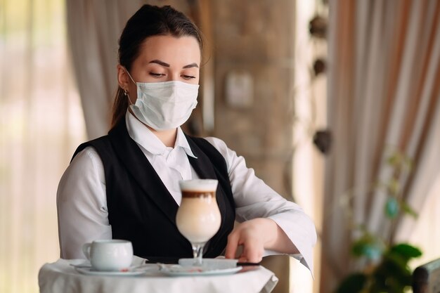 Eine Kellnerin mit europäischem Aussehen in einer medizinischen Maske serviert Latte-Kaffee.