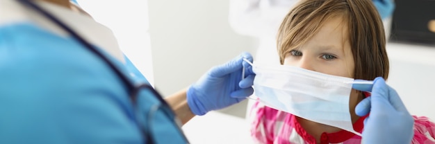Eine Kinderärztin in einer Maske setzt eine Schutzmaske für ein Mädchen auf, ein Kind bei einem Arzt