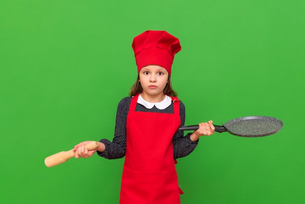 Eine Köchin eines kleinen Mädchens hält ein Nudelholz für Teig und eine Bratpfanne, die mit einer roten Schürze und einer Kochkappe auf einem isolierten grünen Hintergrund bekleidet ist