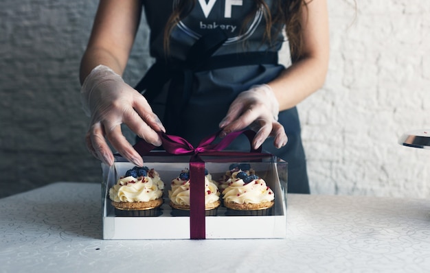 Eine Köchin in einer grauen Schürze packt Cupcakes mit Sahne in eine Geschenkbox, um die Bestellung an den Kunden zu senden. Backen zu Hause.