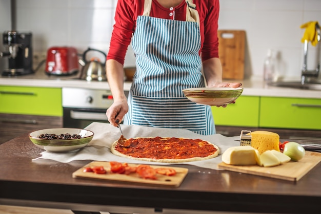 Eine Köchin in einer Schürze gibt einer rohen Pizza Tomatensauce