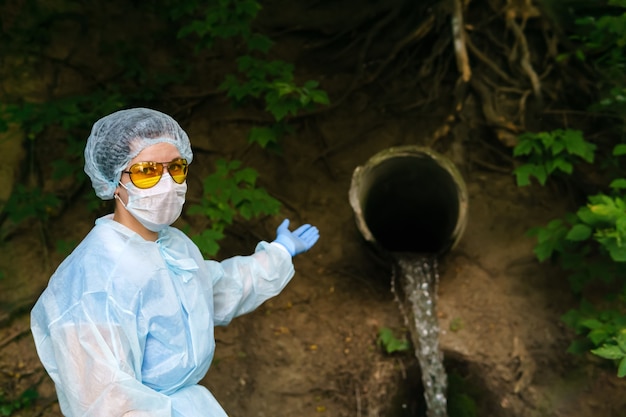 Eine Ökologin oder Epidemiologin in einem Schutzmantel und einer Maske weist auf einen Abwasserkanal hin