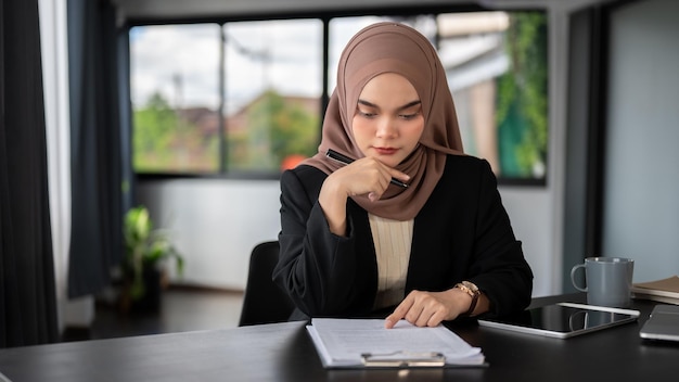 Eine konzentrierte asiatische muslimische Geschäftsfrau, die einen Hijab trägt, prüft Geschäftsdokumente an ihrem Schreibtisch