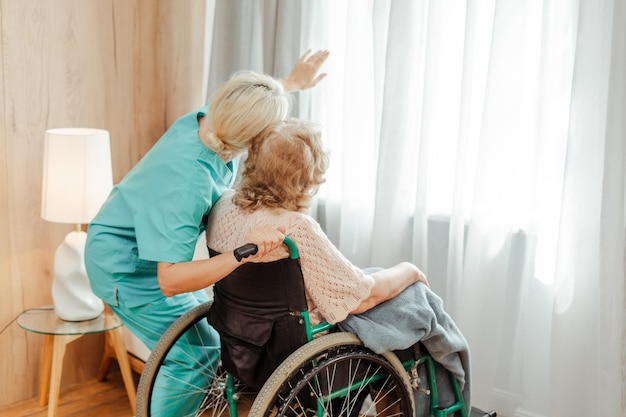 Eine Krankenschwester unterstützt eine ältere Frau im Rollstuhl, die aus dem Fenster schaut