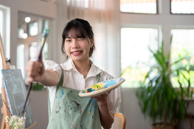 Eine kreative asiatische Künstlerin hält einen Pinsel und eine Farbpalette in der Hand, während sie im Studio steht