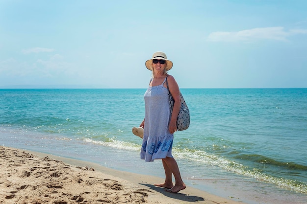 Eine lächelnde ältere Frau in einem Sommerkleid und einem Hut geht mit einer Tasche und Schuhen in der Hand am Strand entlang. Aktiver Lebensstil, Tourismus, Erholung und Reisen