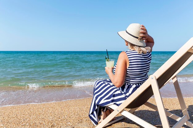 Eine lächelnde ältere Frau sitzt mit einem Getränk in den Händen auf einer Liegestuhl am Meer, eine Senora in einem blau gestreiften Kleid und Hut, aktive Erholung im Resort und Entspannung.