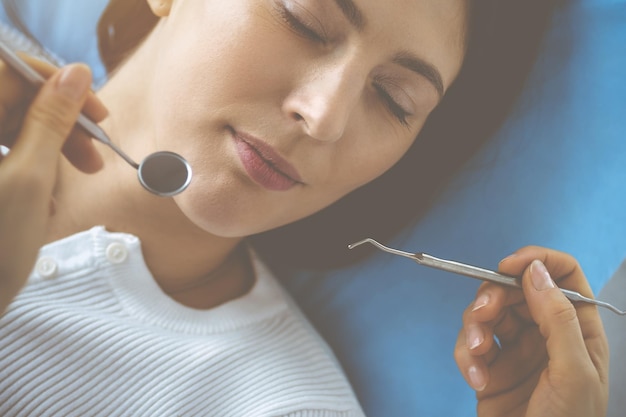 Eine lächelnde brünette Frau wird von einem Zahnarzt in einer Zahnklinik untersucht. Die Hände eines Arztes halten Zahninstrumente in der Nähe des Mundes des Patienten. Gesunde Zähne und Medizinkonzept.