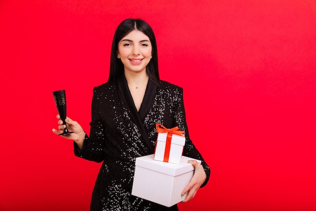 Eine lächelnde Brünette in einem glänzenden, eleganten Kleid hält Schachteln mit Weihnachtsgeschenken und ein Glas Champagner Studioaufnahmen auf rotem Hintergrund