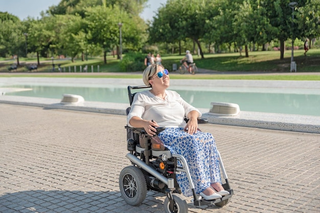 Eine lächelnde Frau im Elektrorollstuhl genießt einen Spaziergang an einem sonnigen Tag im Stadtpark in der Stadt mit Blick auf die Sonne